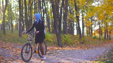 Muslim woman in a hijab and with a backpack with a bicycle goes through a sunny autumn park, sun glare,slow mo