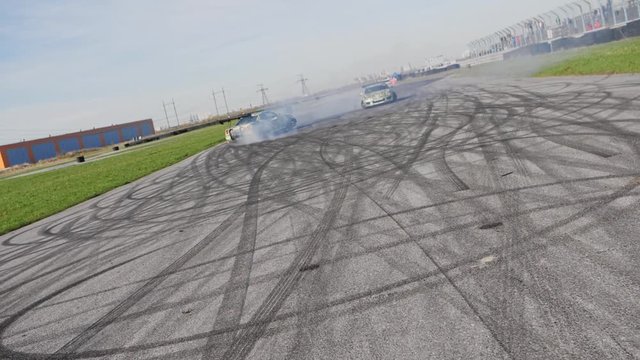 Two green muscle drifting cars on a race track doing a parallel drift around each other with deep tire marks on asphalt, slow motion