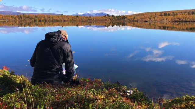 Hiker reading his map sitting by the edge of a crisp clear mountain lake during autumn in Abisko, Lapland, Sweden.