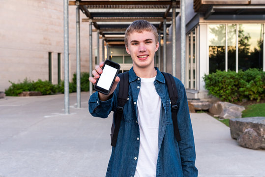 Happy Male Teenager Showing Off His Phone As He Stands In Front Of The Entrance To His High School.