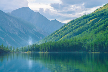 Naklejka na ściany i meble Ghostly mountain lake in highlands at early morning. Beautiful misty mountains reflected in calm clear water surface. Smoke of campfires. Amazing atmospheric foggy landscape of majestic nature.