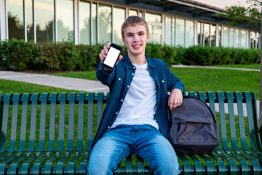 Happy Male Teenager Showing Off His Phone While Sitting On A Bench.