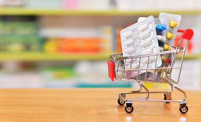 Shopping cart filled with blister packs of medicine on pharmacy drugstore shelves background.Online medical concept.
