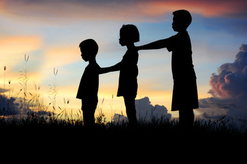 Black silhouette of three children standing shoulder to shoulder. There is a sky at sunset.