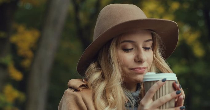 Portrait shot of the charming young blond woman in a hat drinking hot coffee and then smiling to the camera in the park in autumn. Close up.