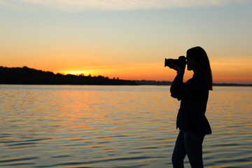 Young female photographer taking photo of riverside sunset with professional camera outdoors