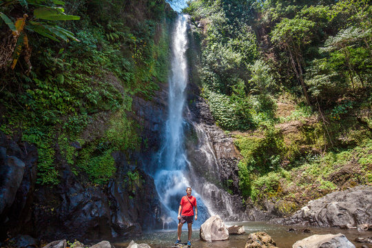Man in red t-shirt standing on the stones in front of Gitgit waterfall among green tropical trees on the Bali island, Indonesia