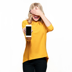 Gorgeous laughing woman showing blank screen mobile phone over white background, covering her eyes...