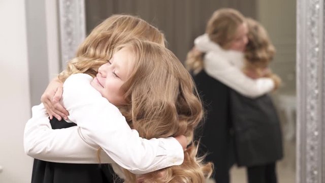 A close up of a charming fair-haired girl standing in the room. She makes a step forward a younger girl and they hug. Their reflection is blurred in the mirror behind them.