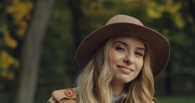 Portrait shot of the beautiful young Caucasian woman in a hat smiling and waving her hand to the camera while sitting in the fall park. Close up.