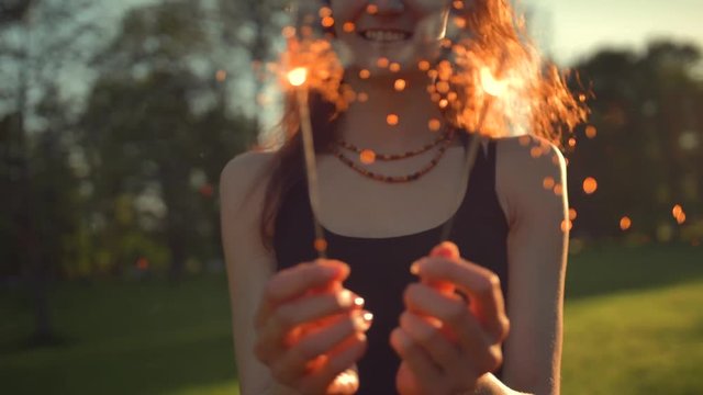 A young woman with a sparkler shake it and dancing in summer evening green park sunset. Bengal light 4k uhd.
