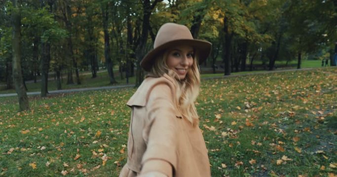 Back view on the young blonde woman in a hat and coat walking the autumn park and leading somebody with her hand tothe camera like follow me, then turning and smiling. Rear.