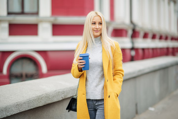 Stylish happy young woman wearing  bright yellow coat. She holds coffee to go. and happily talking on the phone on the street. Portrait of a young cheerful woman at work break