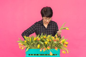 Floristics, holidays and people concept - Middle-aged woman holding box of tulips on a pink background