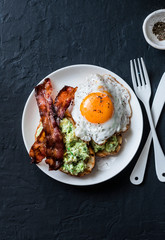 Healthy nutritious breakfast - avocado toast, bacon and fried egg on dark background, top view