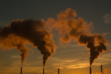 Smoke or gas billowing from industrial smokestack chimneys, lit orange and red by the morning sunrise.