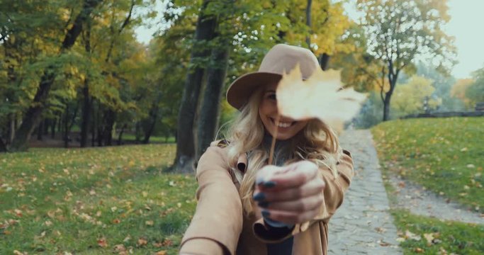 Portrait shot of the blonde cheerful young woman walking the autumn park, playing with leaves and doing follow me to the camera. Close up.