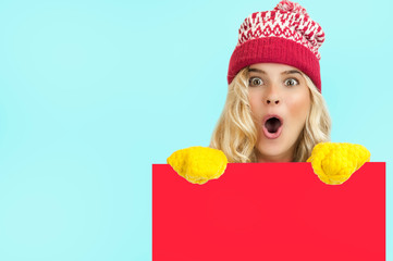 A portrait of a very beautiful bright girl in Christmas clothes posing behind a red panel isolated on a blue background Holding a blank Christmas billboard