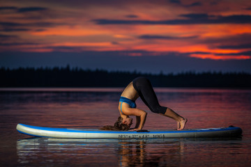Beautiful slim fit girl with long brown hair exercising yoga on paddleboard in the dark, colorful sunset on scenic lake Velke Darko near Zdar nad Sazovou, Czech Republic
