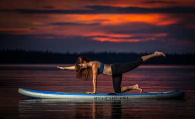 Beautiful slim fit girl with long brown hair exercising yoga on paddleboard in the dark, colorful sunset on scenic lake Velke Darko near Zdar nad Sazovou, Czech Republic