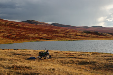 Obraz na płótnie Canvas Motorcycle enduro traveler with suitcases standing alone on sunset above highland steppe shore of lake with dry yellow grass on the background of rocky mountains under clear sky