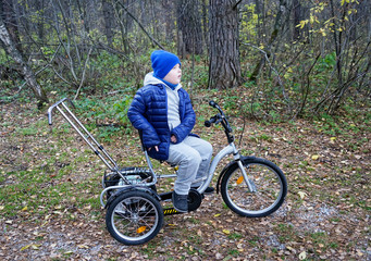 child autistic with disabilities on a tricycle with management for mom, in a blue cap and jacket in the Park for a walk
