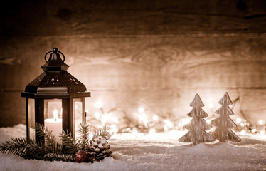 Christmas scene with a lantern, trees, fir branch, snow flakes and blurred lights in front of an illuminated dark wooden board as copy space