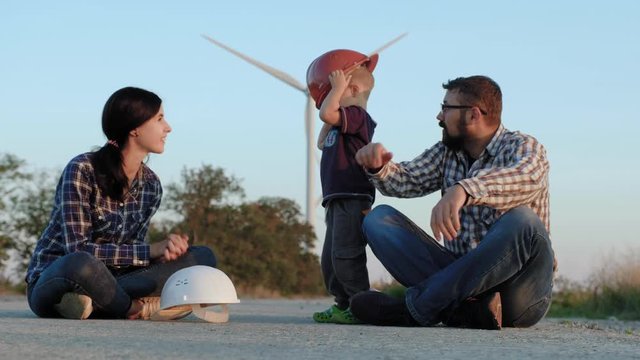 A happy family sits on a road. A child plays with constructor helmets, kisses mother and high-fives father