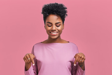 Funny surprised African American female looks with joyful eyes down and indicates as shows something, sees comic things, isolated over pink wall. Positive amazed young woman poses in studio © antianti