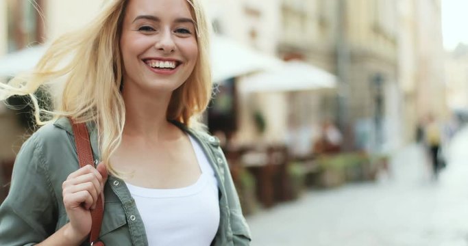 Back view on the blonde young woman walking the town then turning around and smiling cheerfully to the camera. Close up.