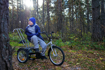 child autistic with disabilities on a tricycle with management for mom, in a blue cap and jacket in the Park for a walk