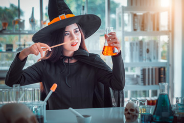 Attractive beautiful Asian woman dressed as a witch using magic wand casting spell to potion in test tube at modern laboratory, Halloween holidays.  