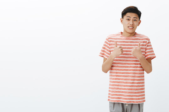 Fine I guess. Portrait of unsure awkward young attractive asian man in striped t-shirt making tight ucertain smile and showing thumbs up gesture as if agree or like idea, posing over gray background