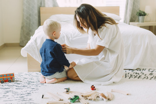 Young beautiful mother buttoning up the shirt of her little son in the bedroom