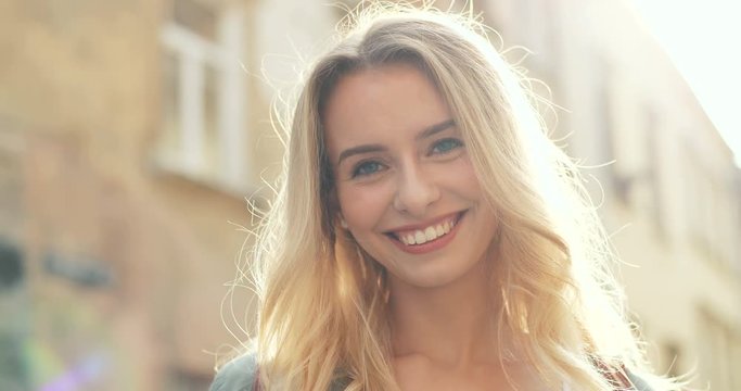 Close up of the charming blond young woman smiling and being happy while standing in the sunlight on the town street. Portrait.