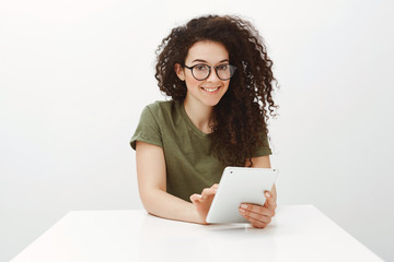 Fototapeta premium Indoor shot of pretty brunette in black glasses, sitting at white table and browsing in network via digital tablet, smiling broadly and feeling carefree while enjoying spending free time in internet