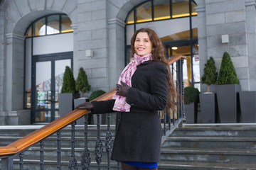 City and people concept - Beautiful and happy woman in black coat and pink scarf standing on stairs