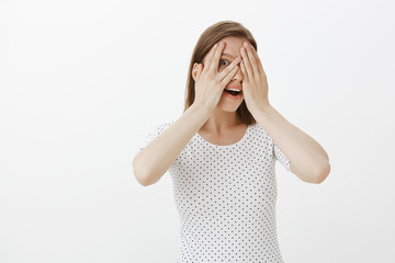 Woman cannot wait, wanting see what waits her, closing eyes with palms, peeking with one eye through fingers, smiling, gasping and gazing with amused and amazed expression, standing over gray wall