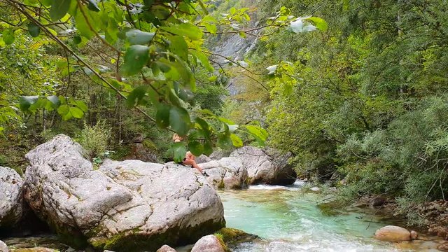 Woman at rapids, UHD tracking shot of a girl behind branches, sitting on a huge rock, looking at the turquoise river, in Trigolov national park, on a sunny summer day, in the Julian alps, Slovenia