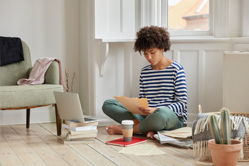 Horizontal shot of administrative manger sits in lotus pose on floor, studies contract, checks documentation, uses laptop computer, drinks takeaway coffee uses scientific literature for writing report