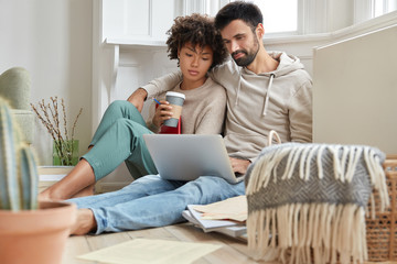 Lovely mixed race couple embrace each other, sits on floor, feel relaxed while watch movie on laptop computer, drink coffee, enjoys domestic atmosphere, connected to wifi. People and leisure