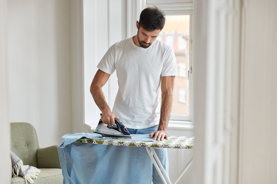 Photo of busy unshaven man irons shirt on ironing board, prepares for formal meeting on business conference, dressed in casual white t shirt, does her domestic duties, stands in spacious room