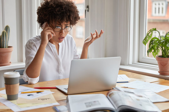 Puzzled shocked indignant female employee shruggs shoulders in frustration, looks at laptop computer, checks database, surrounded with papers, discuss something with colleague, being very emotional