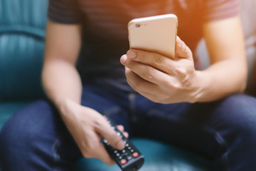 young man using television remote control and mobile smart phone is sitting on a sofa. on or off...