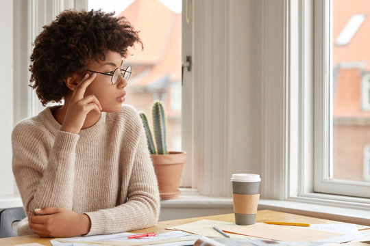 Photo Of Contemplative Woman With Afro Hairstyle, Wears Round Spectacles, Casual Warm Sweater, Thinks About Something While Sits At Desktop, Drink Coffee, Poses Near Window Indoor. Work Concept