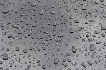 Closeup photograph of raindrops on the hood of a black car. Rain clouds are reflected in the hood.