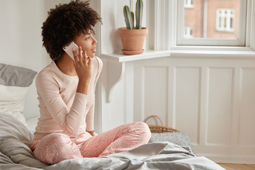 Serious dark skinned girl makes telephone call, satisfied with tariffs and internet connection at home, wears casual nightwear, looks aside, at window, sits on bed, discusses something with friend