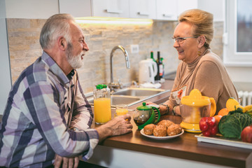 Elderly couple in the kitchen preparing breakfast. They drink coffee or tea and talk