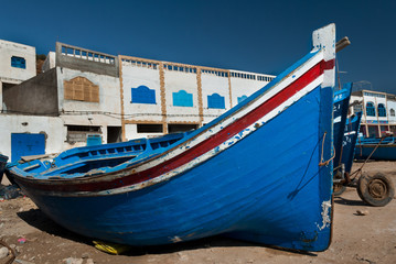 Fototapeta na wymiar A blue boat lays on the ground in front of the Tafelney fishing village buildings