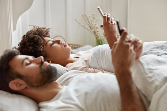 Married mixed race couple lie in bed, being addicted to modern technologies, hold cellulars in hands, play games, surf social networks before falling asleep, pose at bedroom. People, addiction concept
